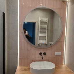 Wykończenia Andrzej Kobylański - Nowoczesna łazienka z różowymi płytkami, okrągłym lustrem z podświetleniem LED i drewnianą szafką pod umywalką. Widoczne czarne baterie i grzejnik drabinkowy.