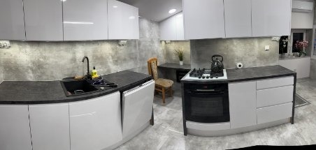 Nowoczesna kuchnia po remoncie: białe, lakierowane szafki, czarny blat, betonowa ściana, oświetlenie LED podszafkowe, stół z krzesłem w tle. Panoramiczny widok.