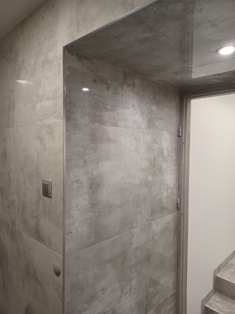 Wykończona łazienka w szarościach: ściany i sufit pokryte płytkami imitującymi beton, widoczny fragment schodów i włącznik światła. Minimalistyczny design.