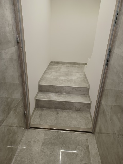 Wykończone schody wewnątrz budynku, wyłożone szarymi płytkami imitującymi kamień, z metalową listwą progową i fragmentem ściany. Widok z dołu schodów.