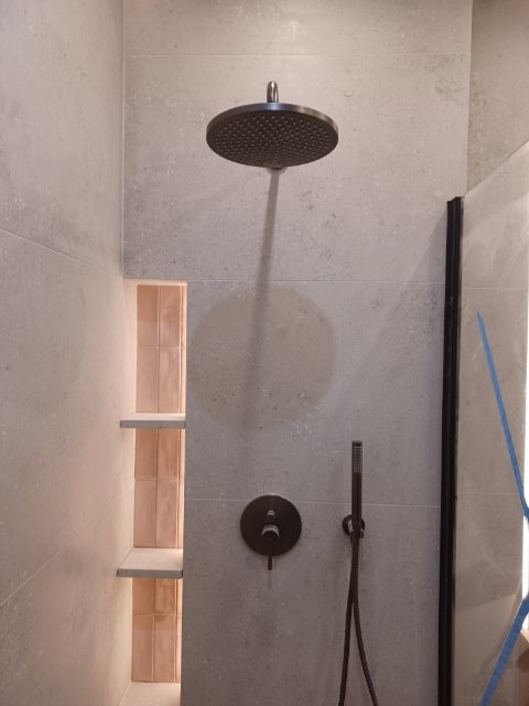 Nowoczesna kabina prysznicowa z deszczownicą i wnęką z półkami wyłożoną różową mozaiką. Minimalistyczny design, szare płytki, czarna armatura.