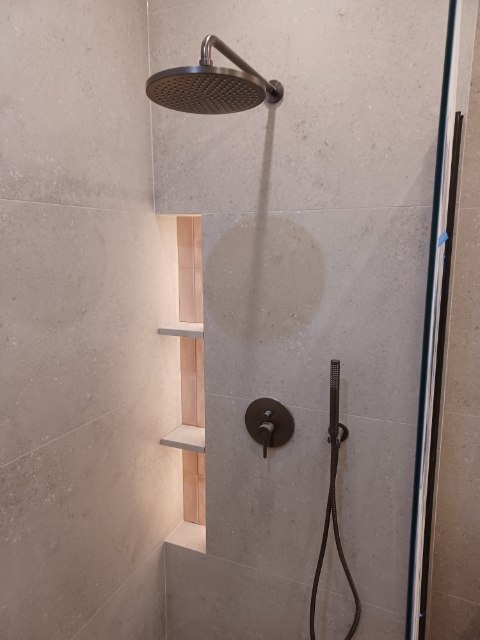 Nowoczesna kabina prysznicowa z szarymi płytkami, deszczownicą i wnęką z półkami podświetlonymi ciepłym światłem. Minimalistyczny design, ciemna armatura.