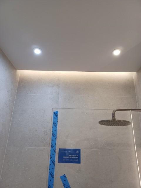 Nowoczesna kabina prysznicowa z deszczownicą i szklaną ścianką, zabezpieczoną folią ochronną. Jasne płytki na ścianach, oświetlenie LED podsufitowe.