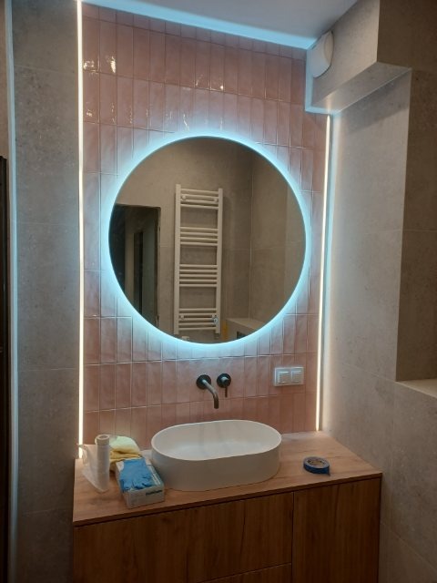 Wykończona łazienka z okrągłym lustrem LED, różowymi płytkami i drewnianą szafką pod umywalką. Widoczne akcesoria glazurnicze i narzędzia pomiarowe.