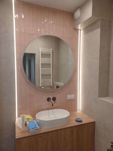 Nowoczesna łazienka z owalnym lustrem i umywalką nablatową. Różowe płytki na ścianie, oświetlenie LED po bokach lustra. Drewniana szafka pod umywalką.