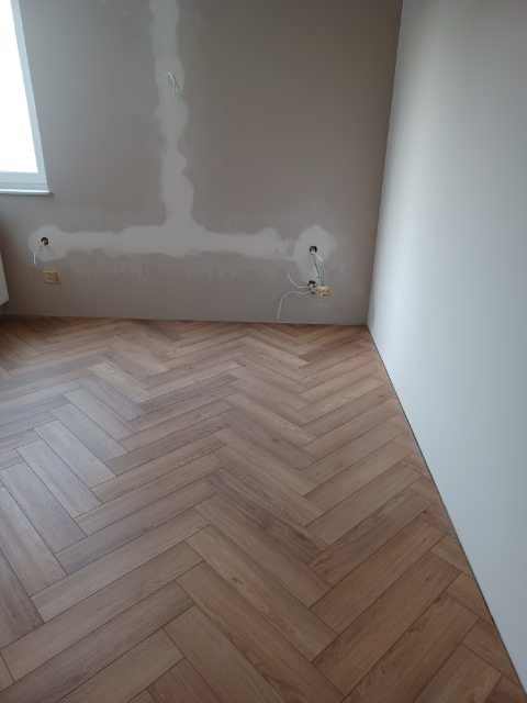 Parkiet w jodełkę, świeżo położony, na tle ściany po szpachlowaniu z widocznymi puszkami elektrycznymi. Jasne wnętrze w trakcie wykańczania.