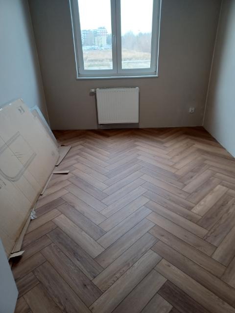 Ukończona podłoga z paneli ułożonych w jodełkę w pokoju z oknem i grzejnikiem. Na podłodze leżą kartony z panelami. Widok na budynki za oknem.
