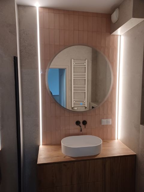 Nowoczesna łazienka z okrągłym lustrem, podświetleniem LED, umywalką nablatową i drewnianą szafką. Różowe płytki na ścianie dodają wnętrzu ciepła i unikalnego charakteru.