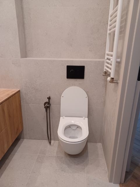 Nowoczesna łazienka z podwieszaną toaletą, bidetką i grzejnikiem drabinkowym. Jasne płytki i drewniana szafka pod umywalką tworzą minimalistyczny, elegancki wystrój.