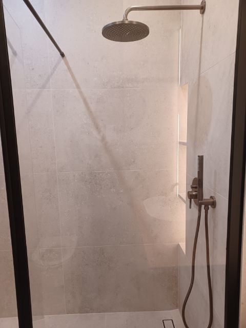 Wykończona kabina prysznicowa z szarymi płytkami, deszczownicą i baterią w kolorze miedzi. Minimalistyczny design z czarną ramą kabiny.