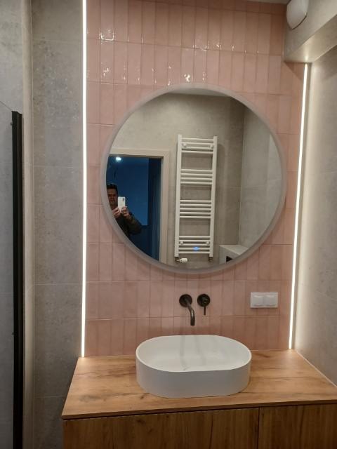 Nowoczesna łazienka z różowymi płytkami, okrągłym lustrem z podświetleniem LED i drewnianą szafką pod umywalką. Widoczne czarne baterie i grzejnik drabinkowy.