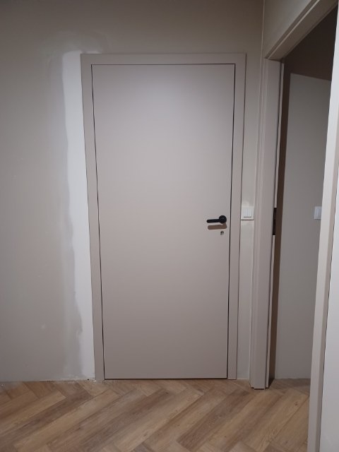 Zamknięte, beżowe drzwi z czarną klamką w nowym futrynie. Ściana częściowo wyszpachlowana, podłoga z paneli ułożonych w jodełkę. Widoczny fragment drugiego pomieszczenia.