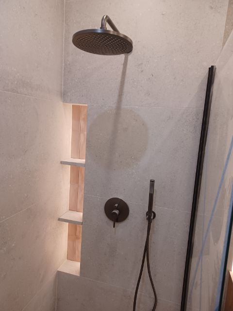 Nowoczesna kabina prysznicowa z deszczownicą i wnęką z półkami, wykończona szarą płytką. Minimalistyczny design, ciemna armatura, szklana ścianka.