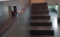 Wykończone schody z ciemnobrązowych płytek, prowadzące w dół, z widoczną ścianą wykończoną szarą płytką i akcesoriami malarskimi na półce.