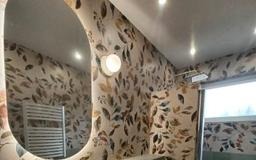 Wykończona łazienka z owalnym lustrem, wzorzystą tapetą w liście i grzejnikiem drabinkowym. Oświetlenie punktowe sufitowe i nad lustrem. Nowoczesny design.