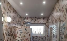 Wnętrze łazienki z tapetą w motywy roślinne, oświetleniem punktowym i grzejnikiem drabinkowym. Widoczne okno z widokiem na drzewa. Nowoczesny design.