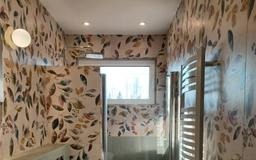 Wąska łazienka z wzorzystą tapetą w liście, grzejnikiem drabinkowym i oknem. Oświetlenie punktowe i kinkiet. Nowoczesny design z naturalnym motywem.