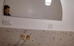 Nowoczesna łazienka: beżowy blat lastryko, złota armatura, owalne lustro z podświetleniem LED i widocznym odbiciem osoby. Minimalistyczne gniazdka elektryczne.