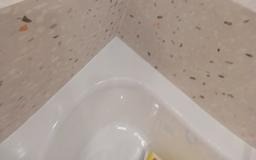 Fragment narożnika łazienki: biała wanna akrylowa i ściana wykończona płytkami imitującymi lastryko w odcieniach beżu i brązu. Minimalistyczny design.