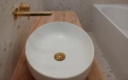 Biała, okrągła umywalka nablatowa z złotą armaturą na drewnianym blacie w nowoczesnej łazience z mozaikową ścianą i fragmentem wanny.