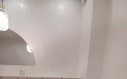 Biała łazienka z lustrem o zaokrąglonym kształcie i lampą wiszącą. Ściany wykończone panelami ściennymi z delikatnym pionowym wzorem. Minimalistyczny design.