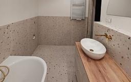 Nowoczesna łazienka z terazzo: beżowe płytki z ciemnymi drobinkami na ścianach i podłodze. Umywalka nablatowa na drewnianym blacie, minimalistyczna armatura.