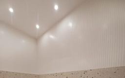 Białe panele ścienne z pionowymi żłobieniami, oświetlone trzema punktowymi lampami sufitowymi. Dolna część ściany wykończona mozaiką w odcieniach beżu i brązu.