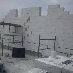 Wznoszenie ściany z białych bloczków na budowie domu, widoczne rusztowanie i zapas materiałów.