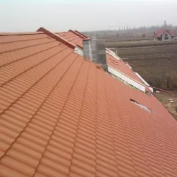 Nowy dach z terakoty z widocznymi elementami konstrukcyjnymi i kominem, w tle krajobraz z domem jednorodzinnym.