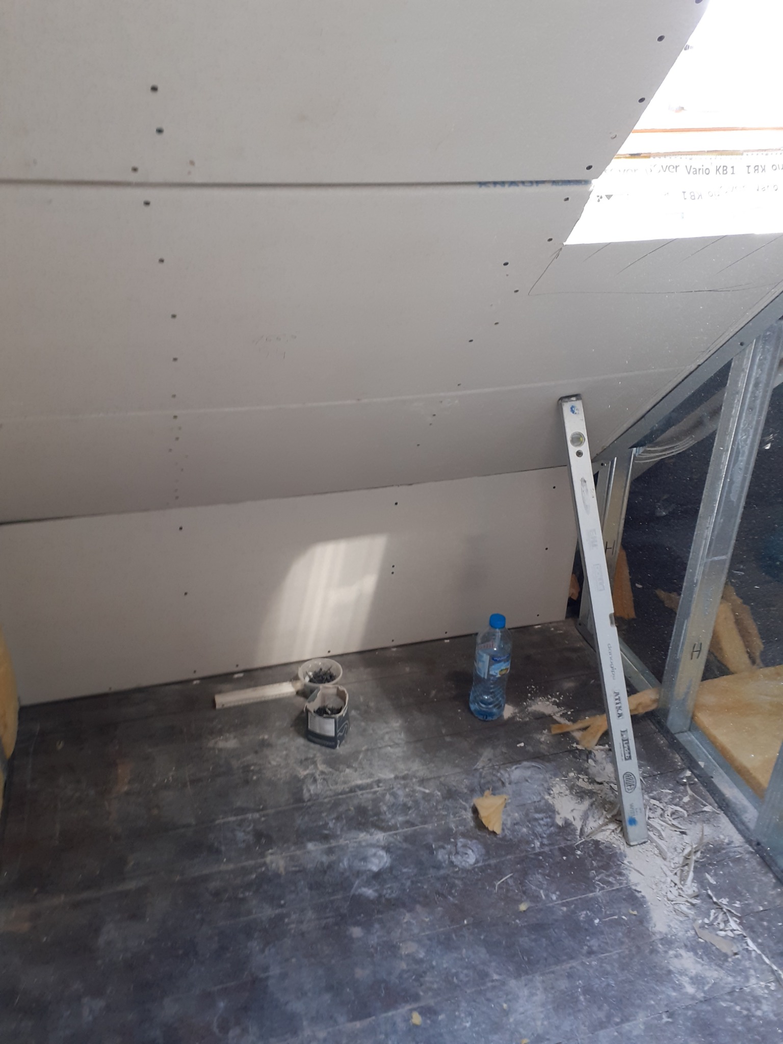 Montaż płyty kartonowo-gipsowej na poddaszu, widoczny poziomica, metalowa konstrukcja, butelka wody i rozsypane resztki materiału na podłodze.