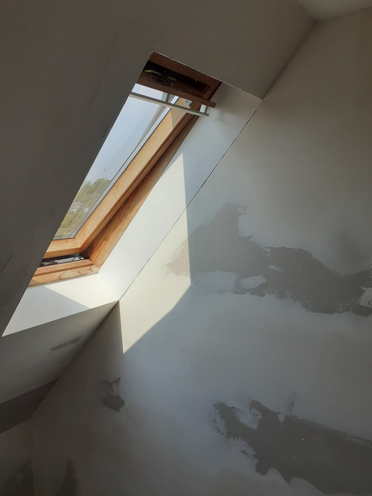 Poddasze z oknem dachowym w trakcie prac wykończeniowych, ściany częściowo pokryte szpachlą, widoczne nierówności i przygotowanie do dalszych prac.