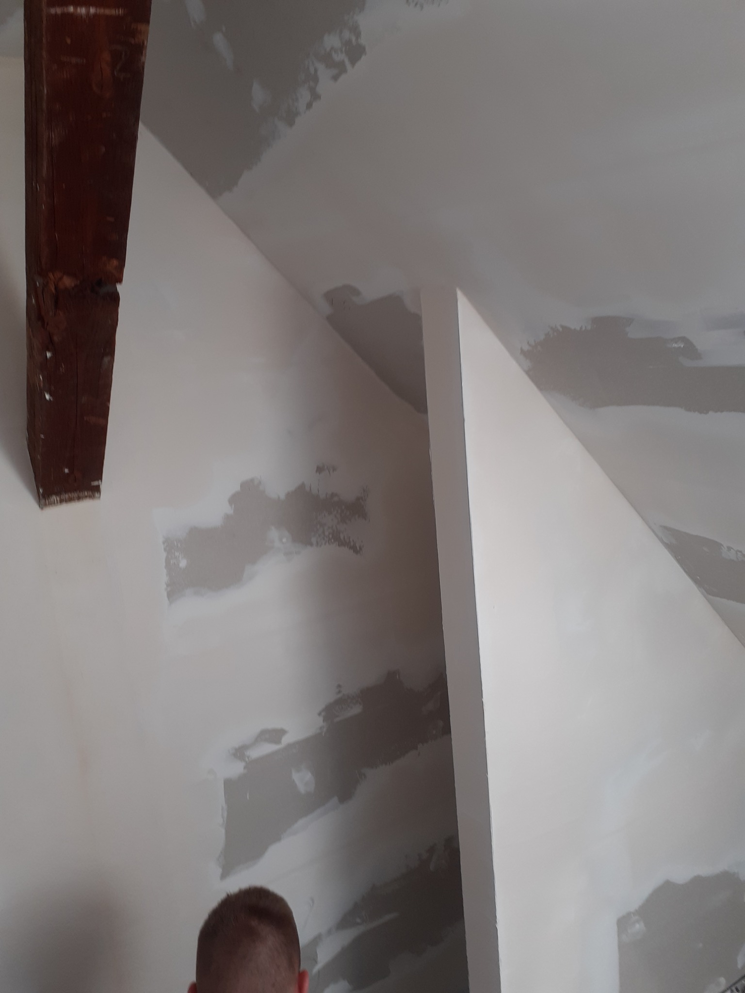 Skośna ściana poddasza z drewnianą belką, częściowo wyszpachlowana, z widocznymi łatami szpachli przed ostatecznym malowaniem.