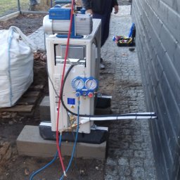 Elektrowniesloneczne.tech - Instalacja klimatyzatora typu split na zewnątrz budynku, z podłączoną pompą próżniową i manometrami, obok czarnej drewnianej ściany i worków z materiałem budowlanym.