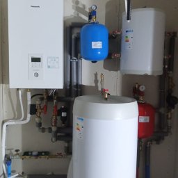 Elektrowniesloneczne.tech - Zestaw urządzeń: biały piec grzewczy Panasonic, niebieski zbiornik wyrównawczy z manometrem, biały i czerwony zbiornik na wodę, połączone rurami w kotłowni z surowymi ścianami z płyt...