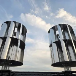 Dwie pionowe turbiny wiatrowe o metalicznej powierzchni na dachu, widoczne na tle błękitnego nieba z chmurami.