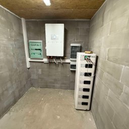 Elektrowniesloneczne.tech - Wnętrze pomieszczenia z zainstalowanym inwerterem fotowoltaicznym, rozdzielnicą elektryczną i modułami magazynu energii na tle surowych, betonowych ścian.