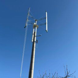 Elektrowniesloneczne.tech - Nisko kątowe ujęcie turbiny wiatrowej z zielonymi akcentami na łopatach, na tle błękitnego nieba. Widoczny betonowy słup i gałęzie drzewa na dole kadru.