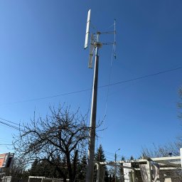 Elektrowniesloneczne.tech - Niska turbina wiatrowa o pionowej osi, zamontowana na posesji, widoczna z dołu na tle błękitnego nieba, z drzewem bez liści w kadrze.