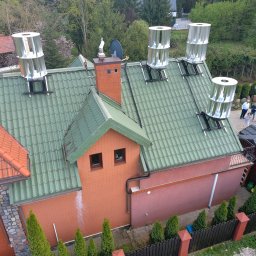 Elektrowniesloneczne.tech - Zielony dach domu z czterema srebrnymi turbinami wiatrowymi Dospel. Widok z góry na instalację, w tle drzewa i grupa ludzi. Czerwona cegła i brązowe okna.