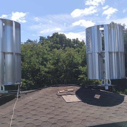 Elektrowniesloneczne.tech - Dwie pionowe turbiny wiatrowe marki Dospel zamontowane na dachu domu z kominem, otoczone zielenią drzew i błękitnym niebem z chmurami.