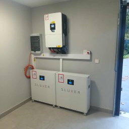 Elektrowniesloneczne.tech - Kompletna instalacja magazynu energii Sluxer w szarym pomieszczeniu. Widoczne dwa moduły na kółkach, inwerter i rozdzielnica. Pomarańczowy kabel łączy elementy.