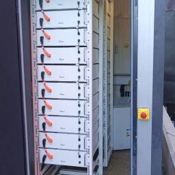 Elektrowniesloneczne.tech - Rząd białych modułów bateryjnych Deye w metalowej szafie, widoczny przez otwarte drzwi. W tle kolejna szafa i instalacja elektryczna. Całość wewnątrz budynku.