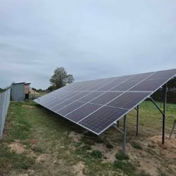 Elektrowniesloneczne.tech - Panel fotowoltaiczny na metalowej konstrukcji, drabina oparta o konstrukcję, ogrodzenie z siatki, budynek w tle, pochmurne niebo. Widok z boku.