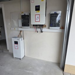 Elektrowniesloneczne.tech - Instalacja fotowoltaiczna w garażu: biały inwerter z oznaczeniami PV, obok rozdzielnice i akumulator na kółkach. Widoczne kable i elementy montażowe.