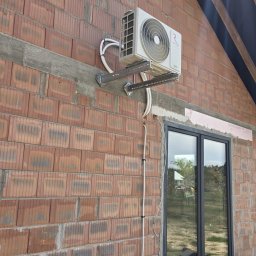 Elektrowniesloneczne.tech - Klimatyzator zamontowany na ceglanej ścianie budynku, widoczne przewody i wsporniki. Świeża elewacja, okno z czarną ramą i skrzynka z napojami w tle.