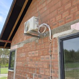 Elektrowniesloneczne.tech - Biały klimatyzator zamontowany na ceglanej ścianie budynku w trakcie budowy, widoczne przewody i elementy montażowe. Okno z czarną ramą w tle.