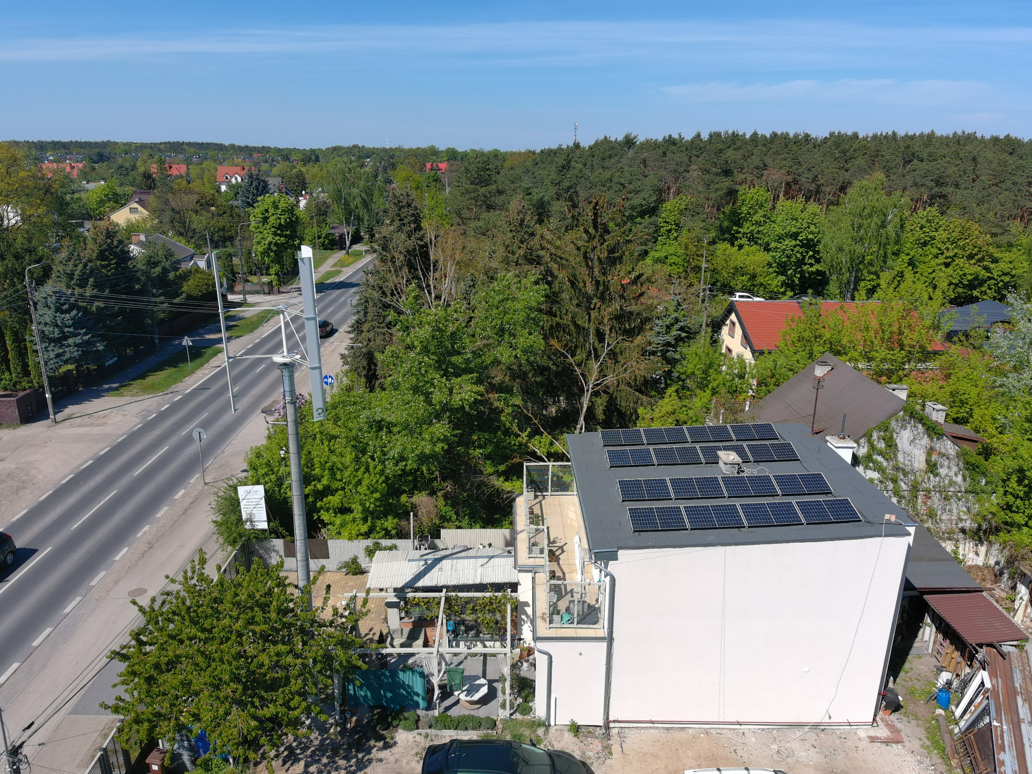 Widok z góry na biały budynek z panelami słonecznymi na dachu, otoczony zielenią i zabudową miejską. Ujęcie z drona, błękitne niebo w tle.