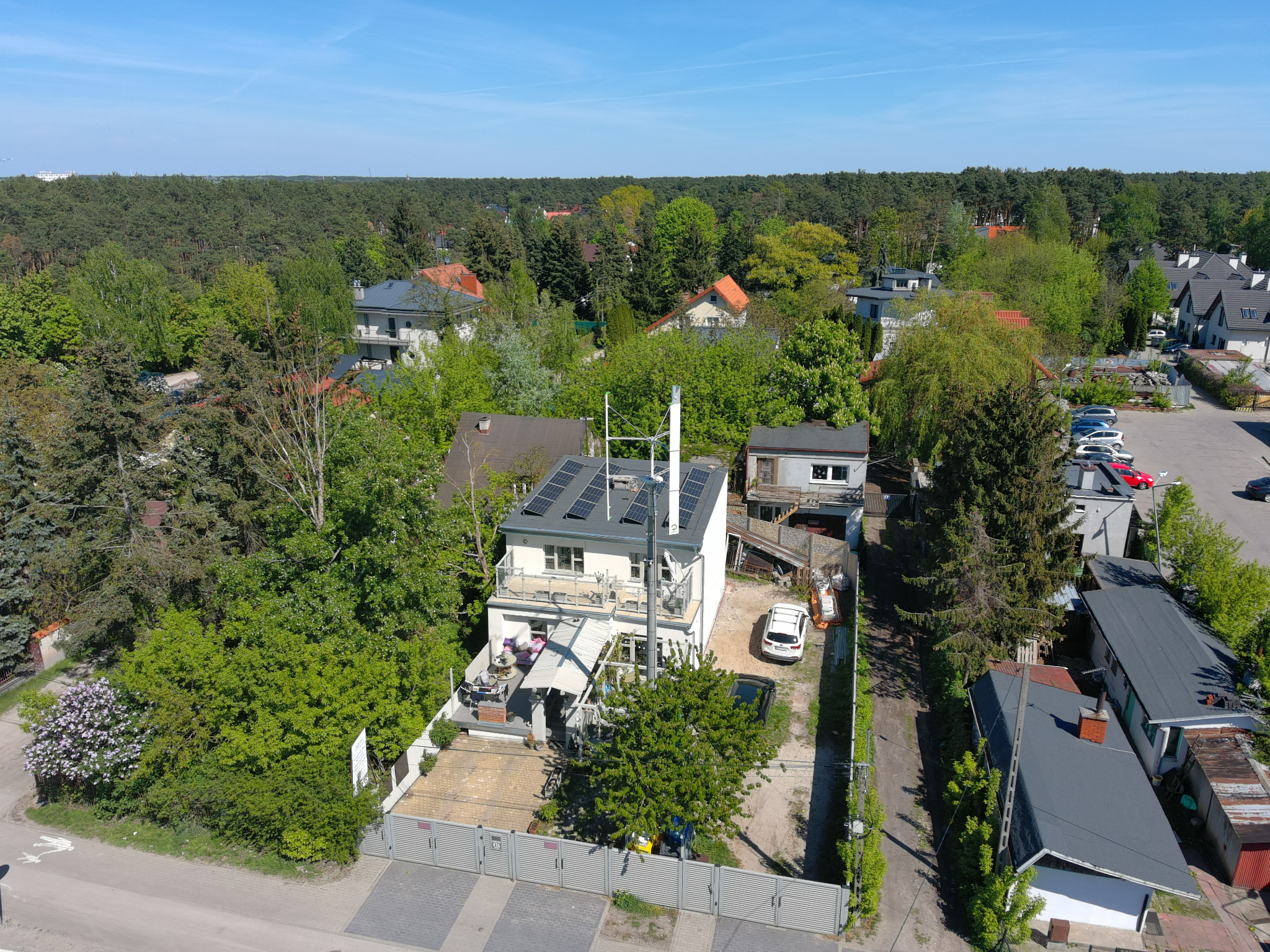 Dom z panelami fotowoltaicznymi na dachu, otoczony zielenią, widziany z lotu ptaka w słoneczny dzień. Biała elewacja, balkon z meblami ogrodowymi.