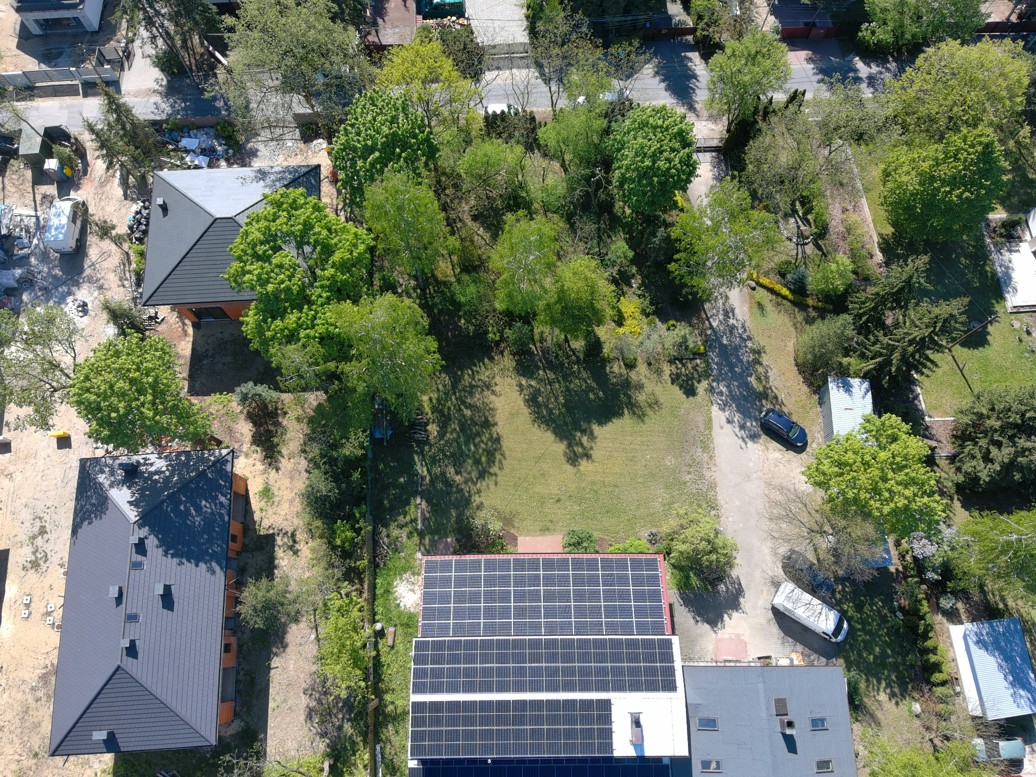 Widok z drona na dom z panelami słonecznymi na dachu, otoczony zielenią i innymi budynkami. Panele zajmują znaczną część dachu.