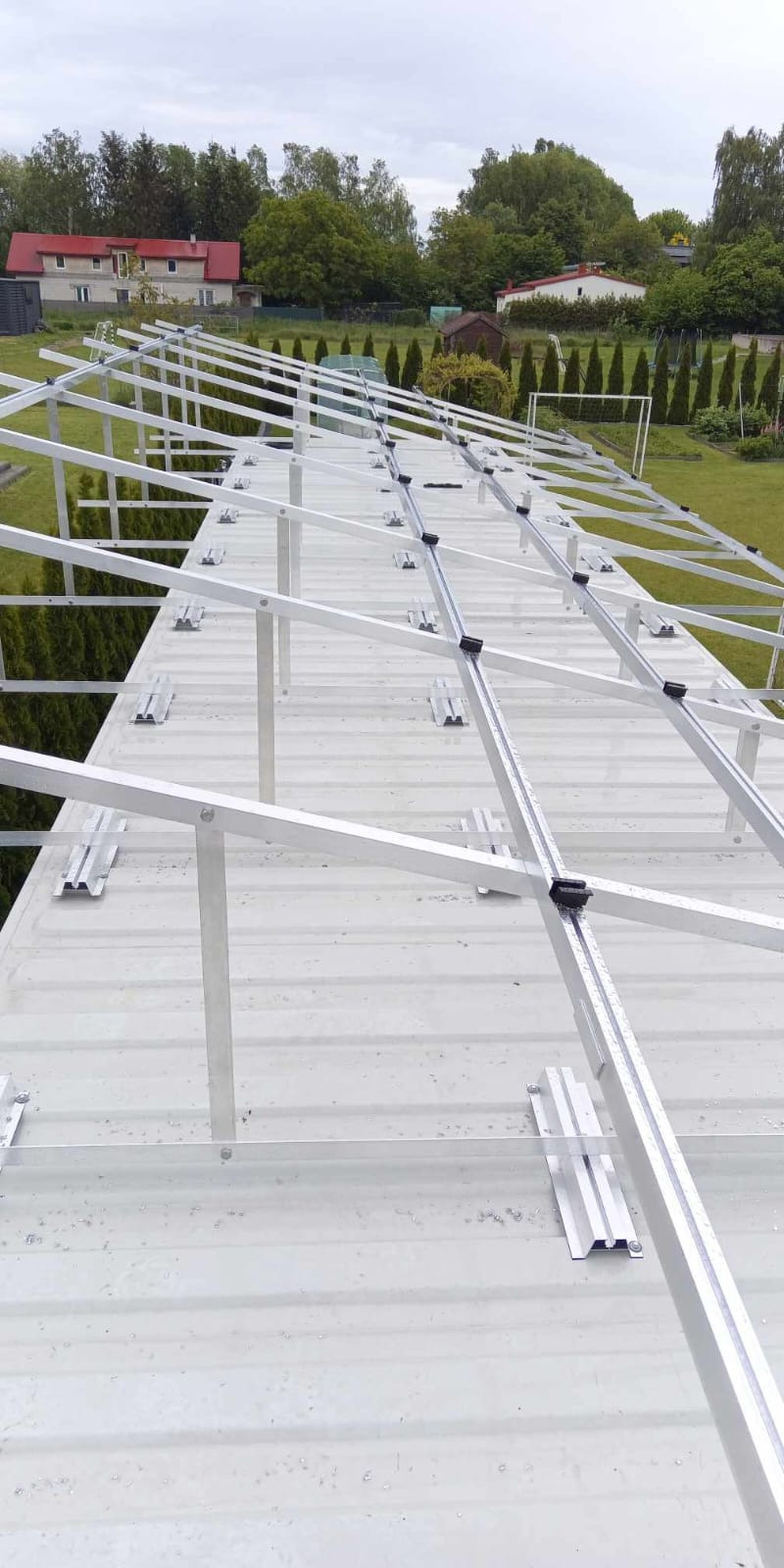 Montaż aluminiowej konstrukcji wsporczej pod panele fotowoltaiczne na blaszanym dachu. Widok z góry, w tle zieleń i zabudowania.